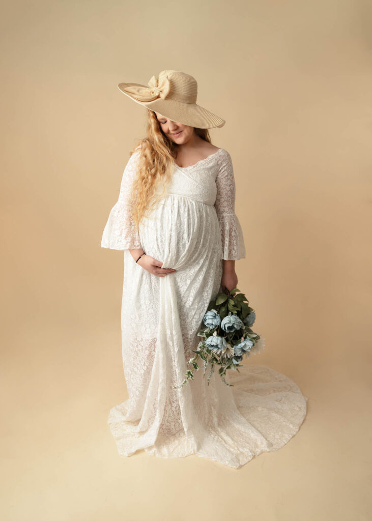 woman in maternity gown with white lace wearing tan hat and holding floral bouquet