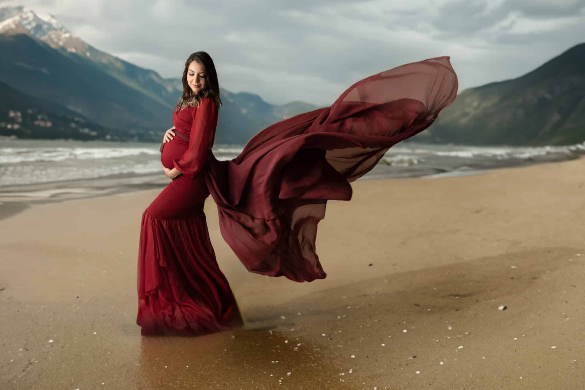 pregnant mom on beach of lake near mountains wearing a red flowing maternity dress