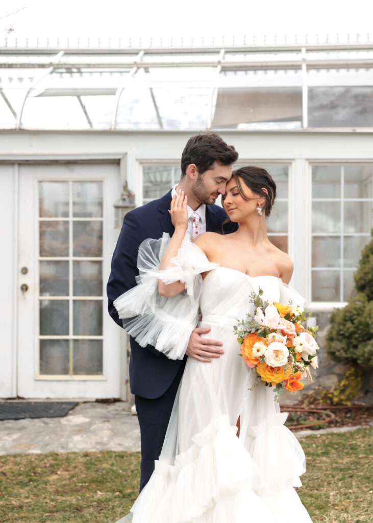 bride and groom snuggling in front of white greenhouse with windows