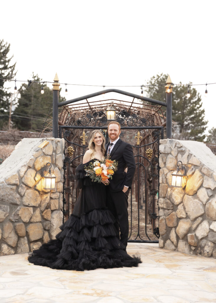 bride and groom in black in front of a garden gate