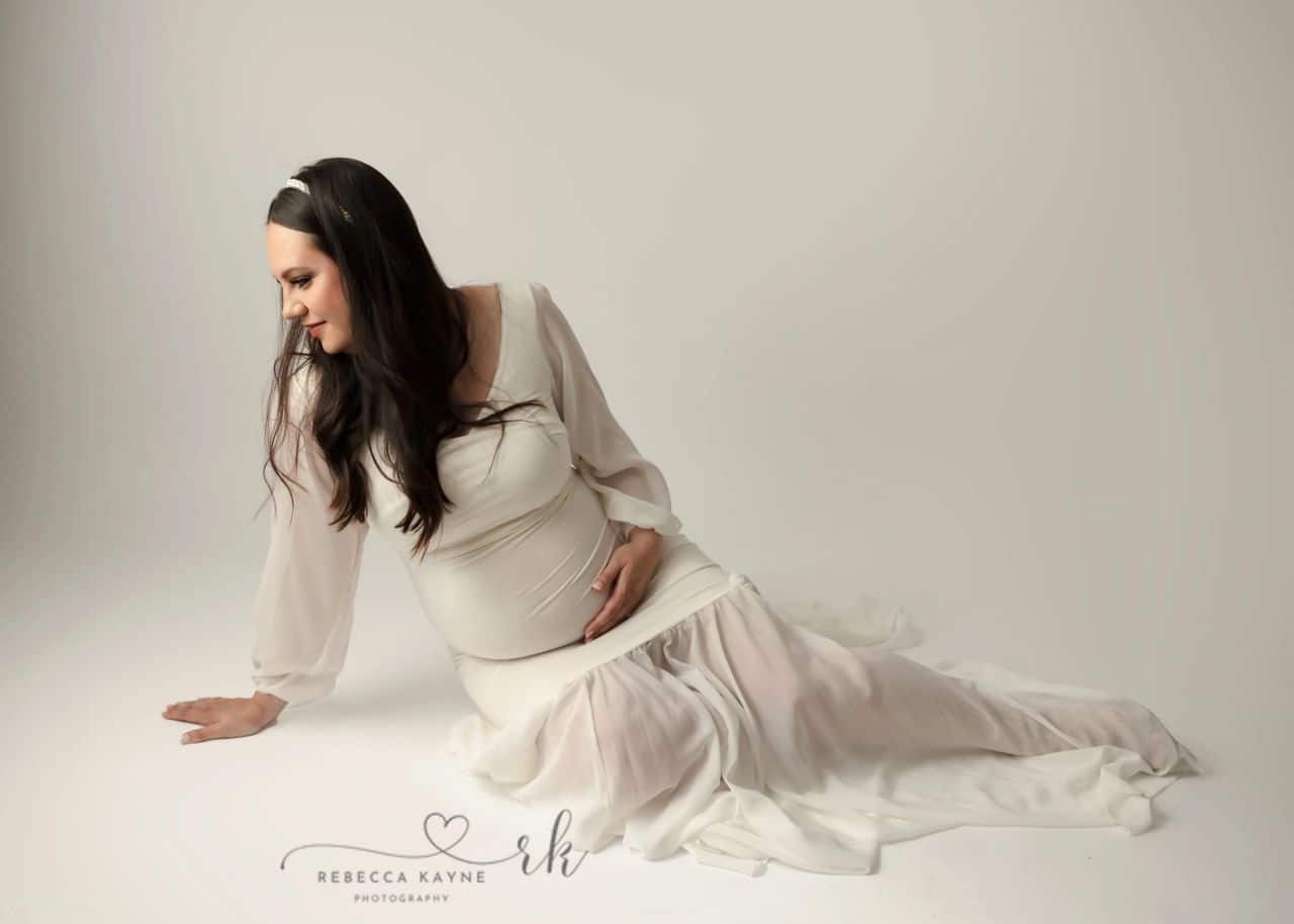 mom in white maternity gown lounging on a white backdrop